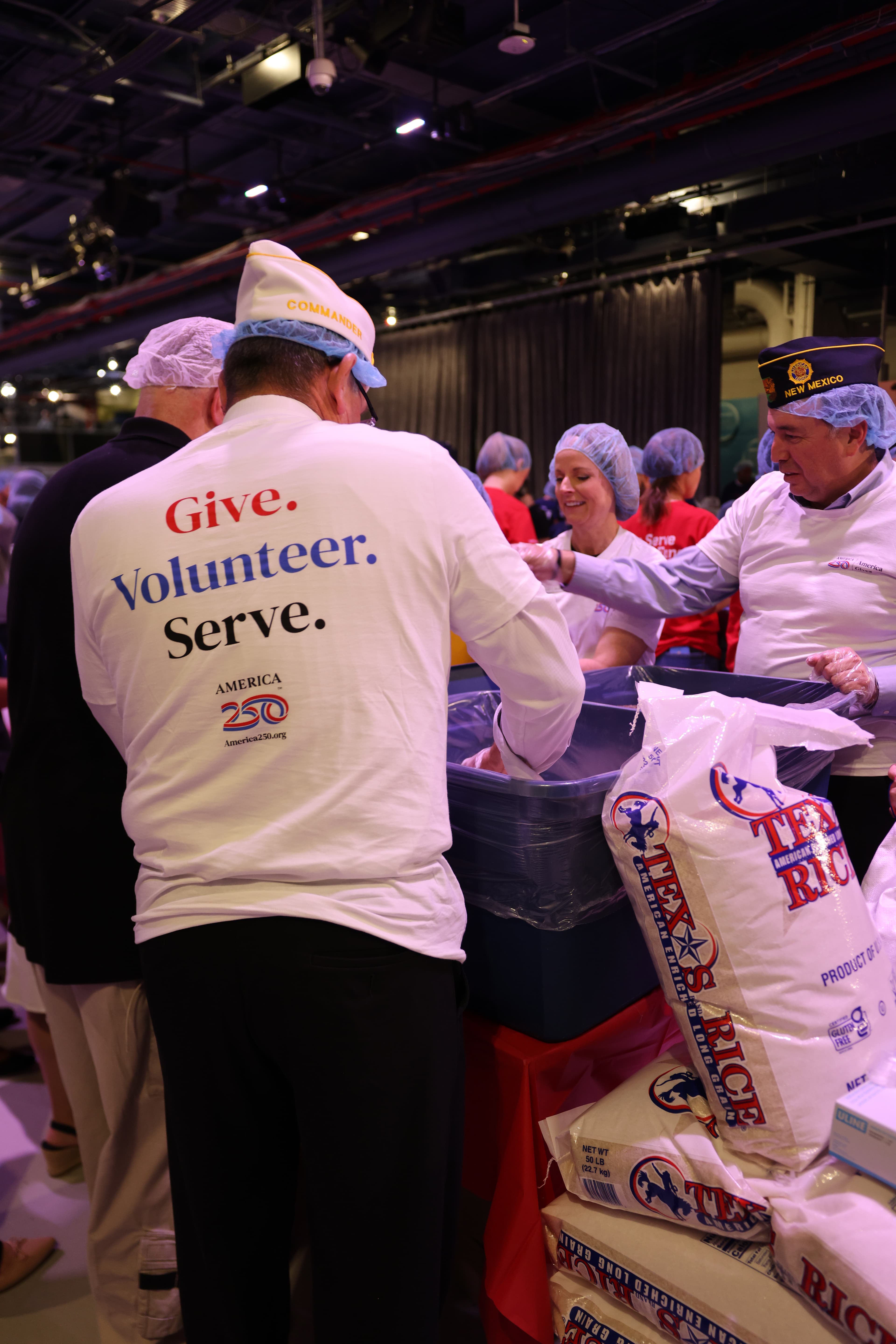 Veterans and volunteers pack food at a service event, wearing shirts that say “Give. Volunteer. Serve.”